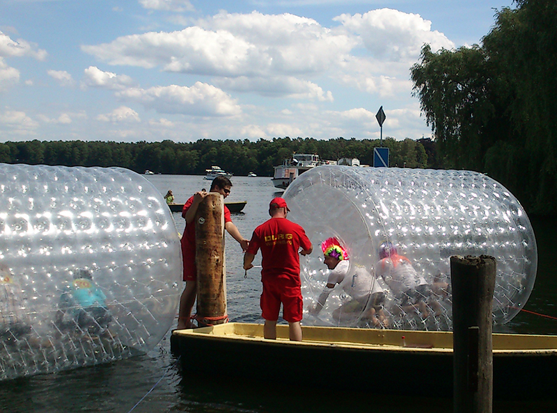 Zwei Personen in transparenten Wasserlaufbällen auf einem See, Betreuer auf dem Steg