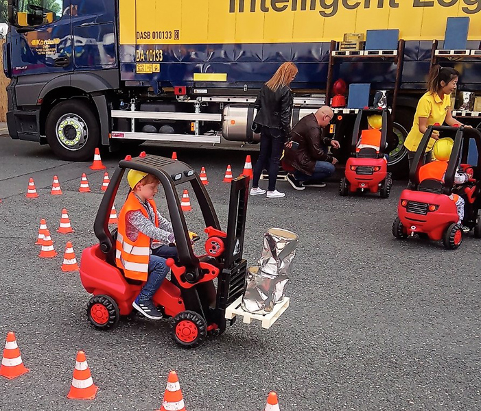 Kind fährt einen Tret-Gabelstapler durch einen Pylonen-Parcours vor einem Lkw