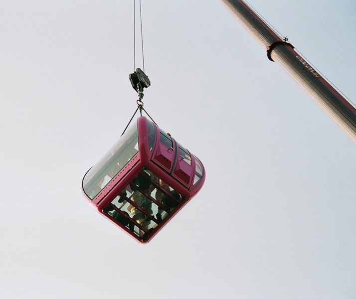 Rote Gondelkabine mit Gästen hängt am Kranarm vor grauem Himmel